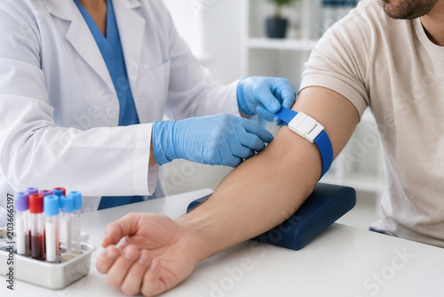 A nurse applies a tourniquet on a patient’s arm to prepare for blood collection. The setting shows medical equipment and blood samples nearby on a table.