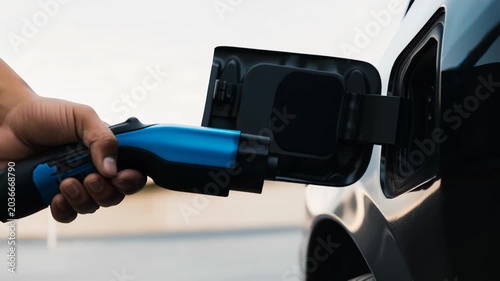 Close up of a person's hand plugging a power supply into a black electric vehicle for charging. Concept of renewable energy and e mobility