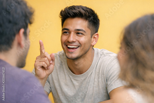 Smiling young adult male in a light blue shirt with dark hair, engaging in conversation, sitting down and pointing.