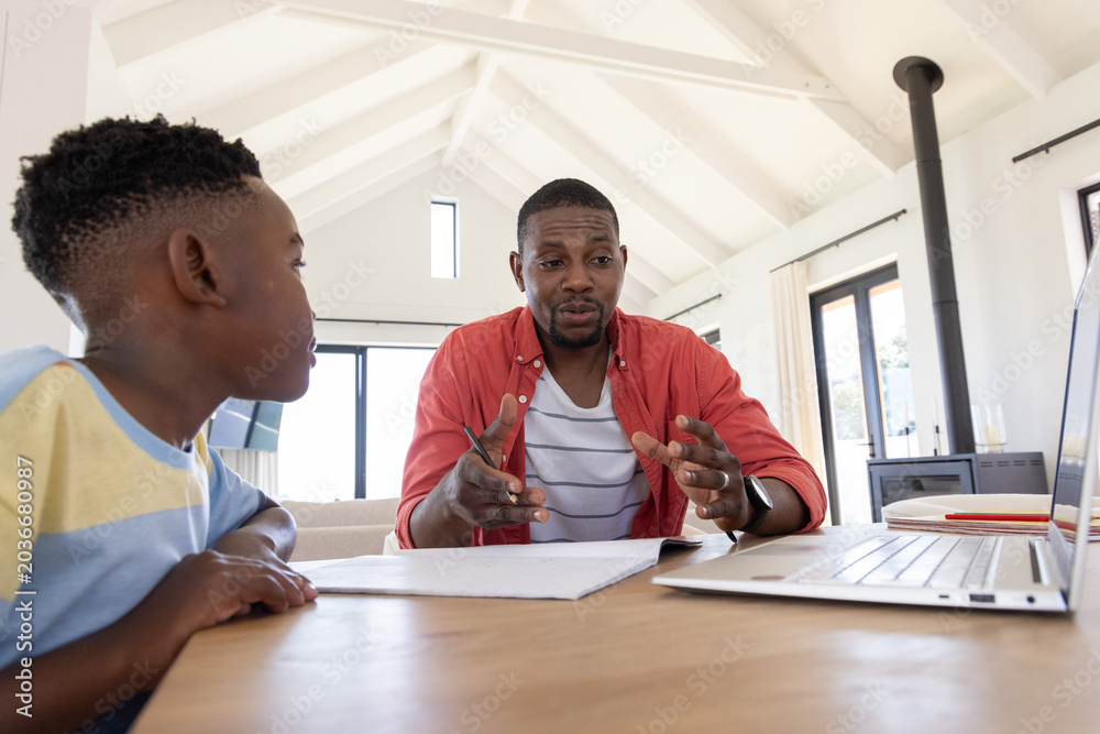 Fototapeta premium African American father and son studying at wooden table at home using laptop and papers