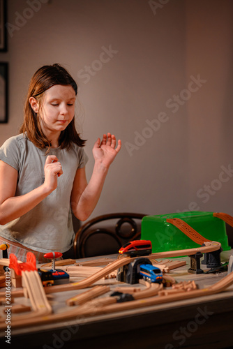 Expressive young girl gesturing while playing with wooden toy train set at table in warm evening light