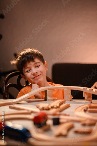 Happy little boy smiling with closed eyes sitting at table with wooden toy railway system