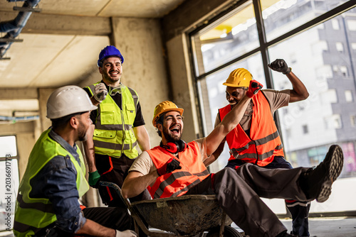 Fotografía Cheerful construction workers playing with a wheelbarrow inside a building site