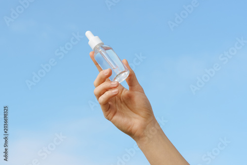 A woman's hand holds a clear glass dropper bottle with serum essential oil against a bright blue sky. Clean, minimalist composition with natural light. Concept for skincare, beauty, wellness, purity