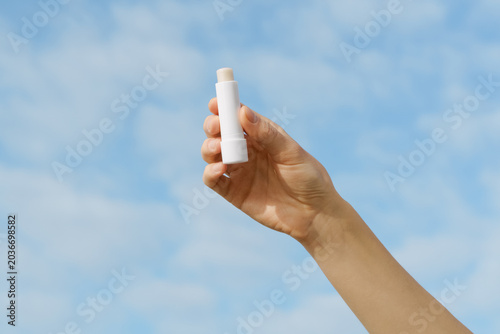 Woman's hand holds a blank white mockup lip balm against a soft blue sky. Minimalist scene with natural light. Concept for skincare, beauty, sun protection, cosmetic mockup, and self care outdoors