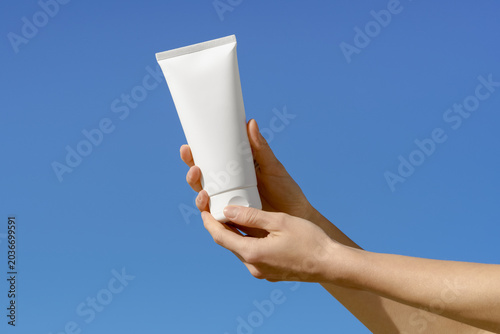 Female hands hold a blank white mockup cosmetic tube against a clear blue sky. Bright natural sunlight creates a clean, minimalist look. Ideal for skincare, lotion, or sunscreen product mockups