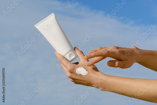 Woman's hands applying white cream from a blank cosmetic mockup tube against a blue sky background. Minimalist skincare concept with natural daylight. For sun protection, beauty, and wellness products