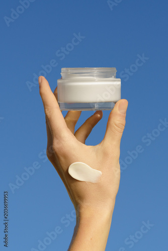 Hand holds a moisturizer mockup jar with a cream swatch on skin. Minimalist product shot with bright daylight against a blue sky. Concept for skincare, beauty, self care, and sun protection