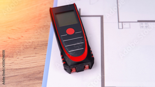 Red laser measure rests on blueprint paper. Black and red colors create sharp contrast. Screen shows measurement numbers clearly. Blue lines mark building plans