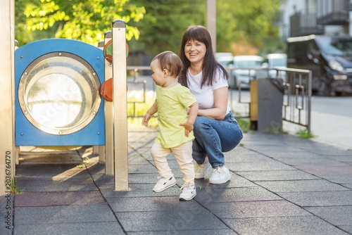 Mother watching toddler explore playground equipment outdoors in summer