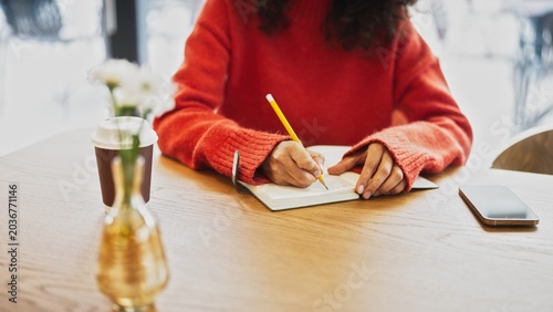 Close up of woman in red sweater writing notes with pencil in notebook at cafe table, takeaway coffee cup nearby. Female with curly hair studying or journaling indoor, smartphone placed on wooden desk