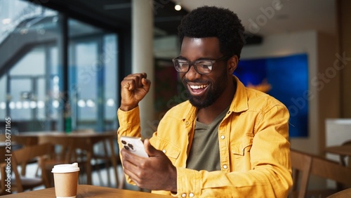 Excited African American man wearing glasses laughing while checking smartphone in cafe. Male smiling with joy looking at phone screen, celebrating success with coffee cup. Victory or good news.