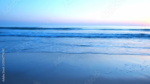 Gentle ocean waves lap onto a serene beach at dawn. The soft pastel sky reflects beautifully on the wet sand.