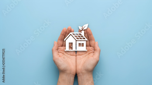 Woman holding paper house with solar panel and leaf growing from roof. Concept of eco friendly home, sustainability and green living. Renewable energy solution for modern estate.