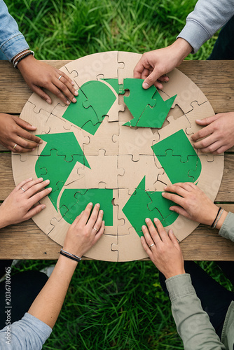 Diverse group of people assembling cardboard puzzle with recycling symbol. Concept of ecology, sustainability and environment protection. Teamwork for nature and earth conservation.