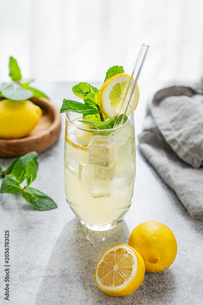 Fototapeta premium A summer drink with lemon, mint, and ice in a glass against a sunny window background.