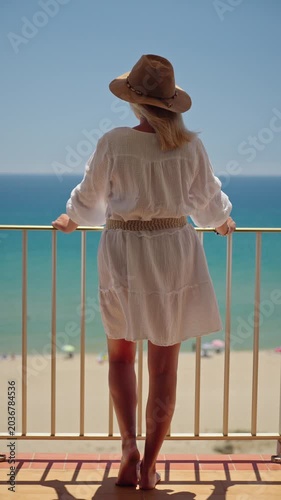 Woman in white dress and straw hat standing on balcony and looking at turquoise sea during relaxing summer beach vacation