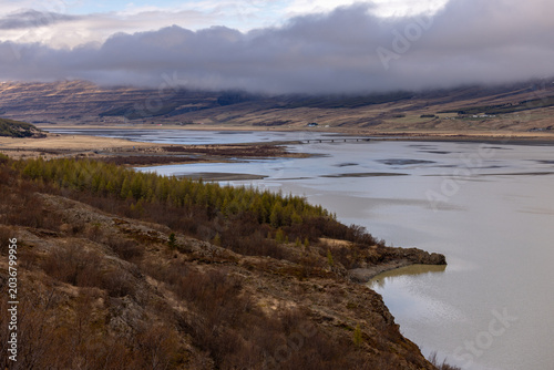 Fotografija Paysage naturel islandais montrant une large rivière, des zones humides et des collines recouvertes de végétation sous un ciel chargé de nuages, illustrant la beauté sauvage du nord
