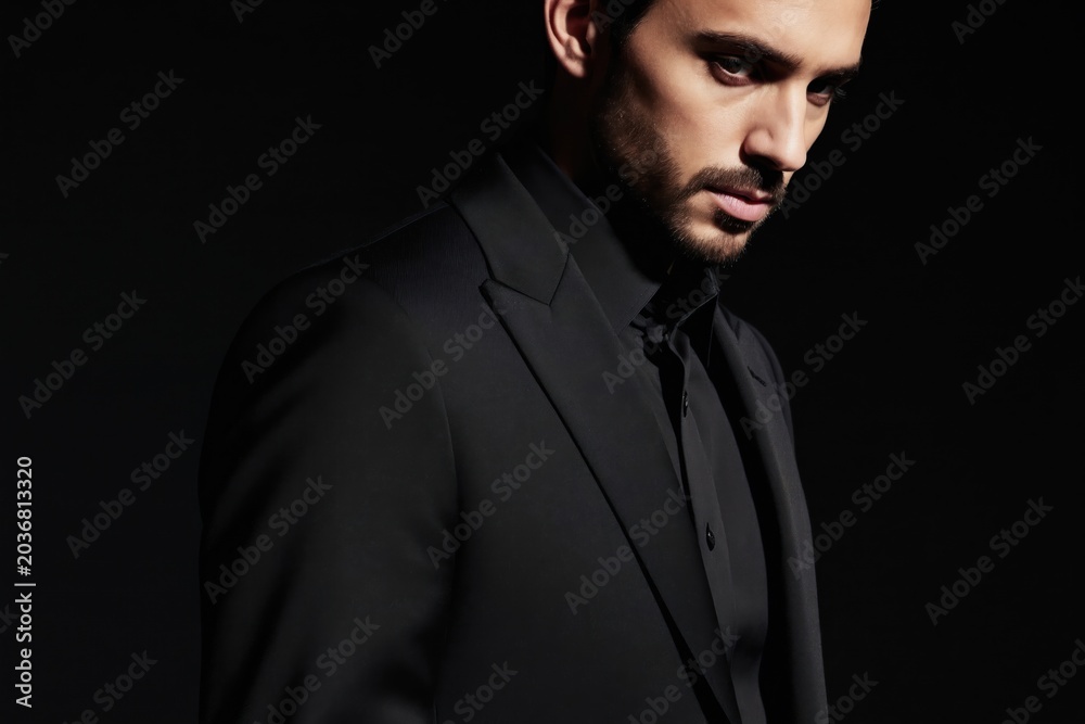 Fototapeta premium Man in black suit poses. Serious look, short brown hair, neat beard. Dark background accentuates sharp features. Pro male portrait. Clean look.