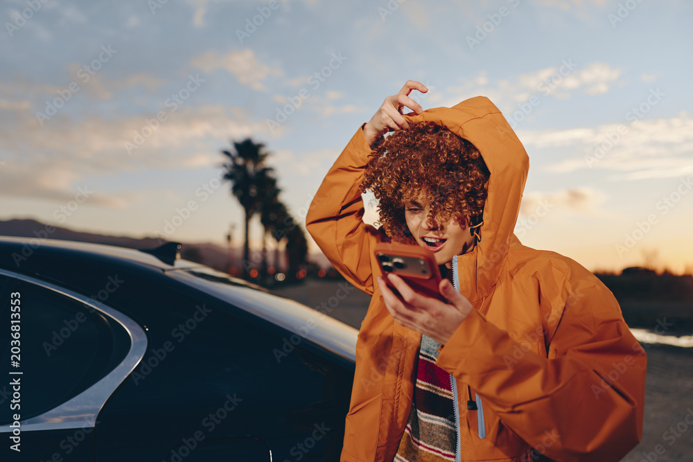 Obraz premium Woman lifestyle portrait, curly hair under hooded orange jacket, smiling, holding phone near car at sunset outdoors, palm trees, warm evening light, casual travel, communication, city road