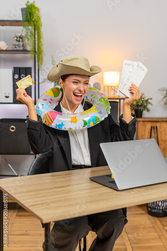 Young businesswoman at home office completes ticket purchase on laptop smiling after payment approval. Freelancer girl at table closes laptop wears inflatable ring dreaming about upcoming vacation fun