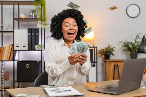 Photography African American businesswoman at home office laughs tossing dollar bills skyward celebrating bonus victory