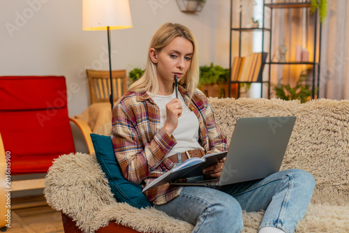 Young adult woman writes in notebook while focusing on laptop screen on home sofa, calculating expenses. Blonde girl manages financial records with care, staying organized and concentrated on task.