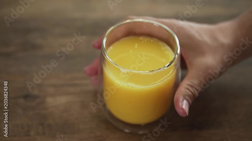 Close-up of a hand gently placing a glass of fresh orange juice onto a wooden table. The scene emphasizes healthy lifestyle, natural vitamin-rich beverage, and morning freshness concept.