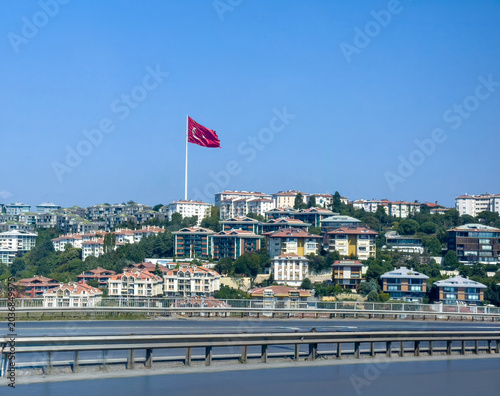 Istanbul, Turkey - July 20, 2024: City views. Giant Turkish flag above neighborhood modern multi-story houses on the north side of the Istanbul ring road 0-1 leading to a long suspension bridge connec