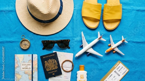 Flat lay of travel vacation essentials on blue background, including straw hat, yellow sandals, passport, sunglasses, and toy airplanes. Concept of summer holiday planning and air travel.
