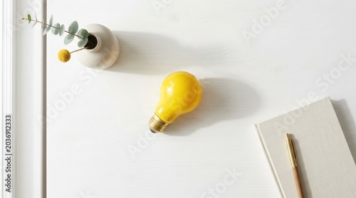Yellow light bulb on white desk with notebook and pencil nearby
