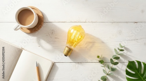 A yellow light bulb sits on a white wooden desk next to a cup of coffee and an open notebook with a pencil.