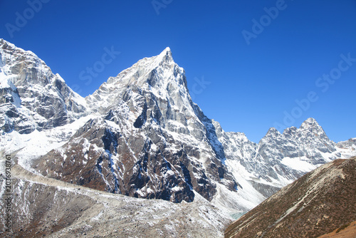 Majestic snow Himalaya mountains in Nepal under a clear blue sky, showcasing rugged peaks and rocky terrain, with a foreground of brown earth and scattered stones visible in the landscape