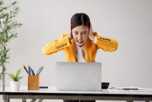 Billede på lærred Young asian businesswoman grimacing and covering her ears at her desk, overwhelm