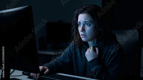 Terrified Young Woman Staring at Computer Monitor in Dark Room, Blue Screen Glow Reflecting on Anxious Face, Cyber Bullying and Horror Concept