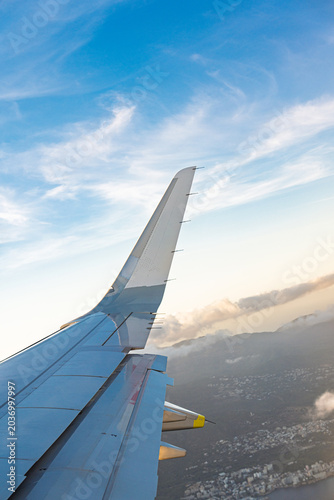 фотографія View from an airplane window showing an aircraft wing over Corfu, Greece, set against a vivid blue sky with scattered clouds, conveying the beauty of travel and the liberating