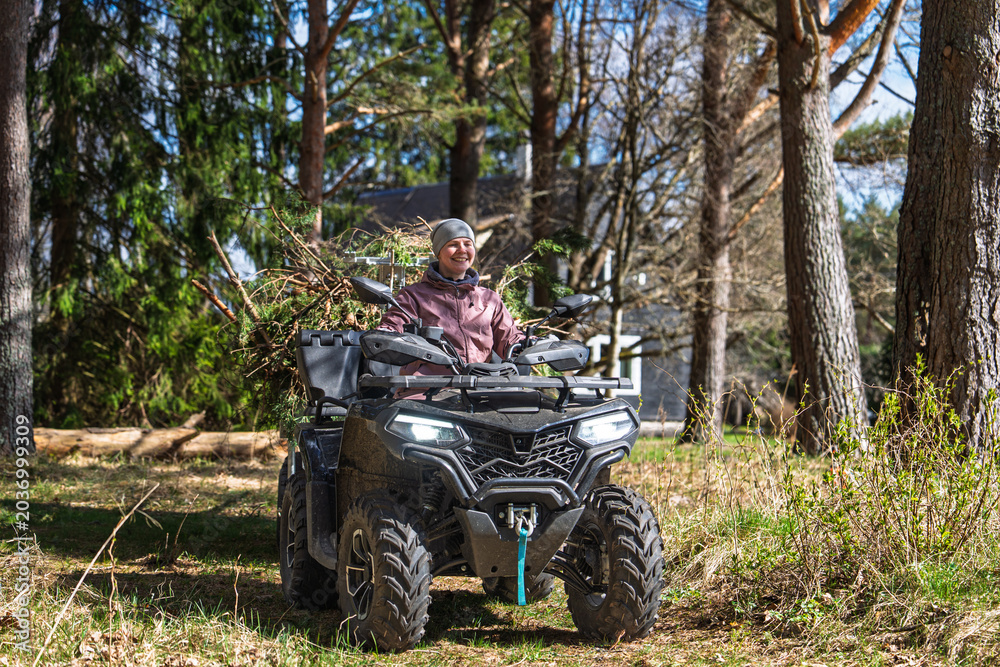 Fototapeta premium A smiling woman is riding an ATV through a forest in Estonia, enjoying the outdoor adventure and exploring the woodland. The all-terrain vehicle is carrying branches for yard.