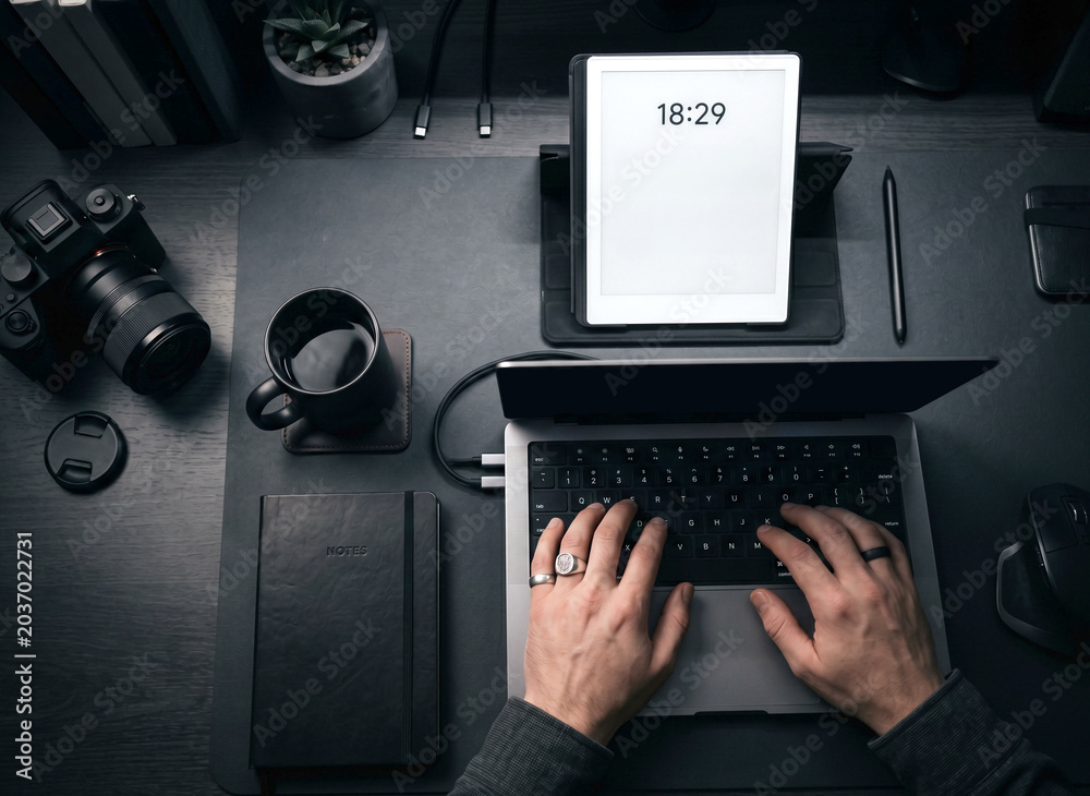 Fototapeta premium Top down view of male hands typing on laptop keyboard on a dark moody workspace with digital tablet mockup, camera, notebook, and coffee.