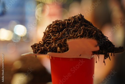 Close up of a creative Thai street food beverage, featuring an iced cocoa drink served in a plastic cup, piled high with a mountain of finely shredded dark chocolate ribbons at a night market stall.