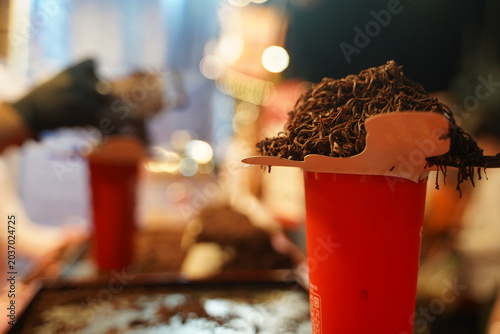 Close up of a creative Thai street food beverage, featuring an iced cocoa drink served in a plastic cup, piled high with a mountain of finely shredded dark chocolate ribbons at a night market stall.