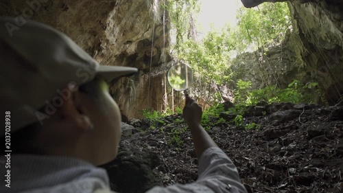 Behind the shoulder view of an Asian woman explorer holding a magnifying glass towards a cave skylight.