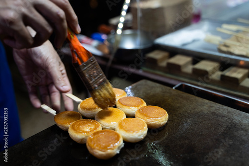 Close up of a street vendor's hand using a brush to oil a hot iron pan while cooking traditional Thai sweet pandan rice cakes (Khanom Krok Singapore) at a night market.