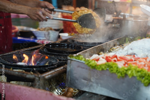 Quadro em tela Close up of street vendor hands using metal spatulas to flip a large crispy mussel omelet (Hoi Tod) amidst steam and smoke on a hot iron skillet at a night market stall