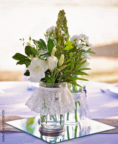 White floral arrangement displayed in lace wrapped glass jars on a mirrored wedding reception table with soft natural light.