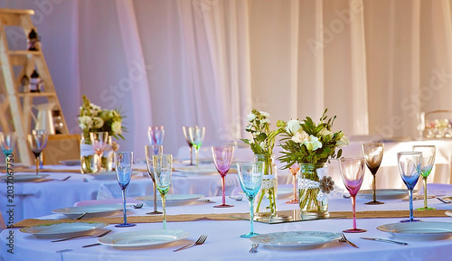 Elegant wedding reception tables decorated with colorful stemware, floral centerpieces, and soft romantic lighting beneath flowing white drapery.