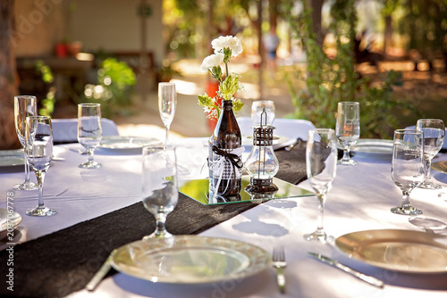 Rustic outdoor wedding table setting with floral bottle centerpiece, lantern decoration, and elegant glassware in warm natural sunlight.