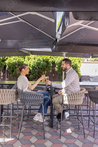 Young man and woman clinking glasses while sitting on high bar stools at an outdoor terrace cafe