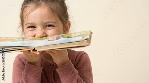 Playful Young Girl Peeking Over Open Book on Beige Background
