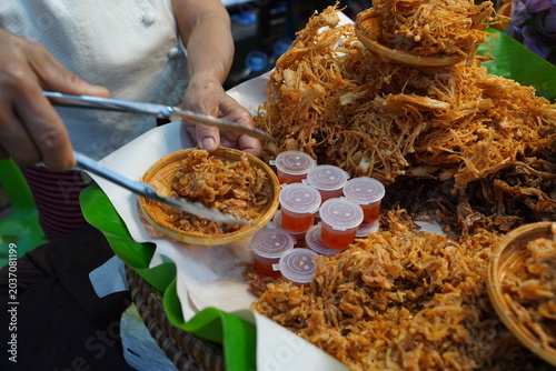 Close up of street vendor hand using tongs to pack crispy shredded sweet pork (Moo Foy), displayed with tiny sweet chili sauce cups at a market stall.