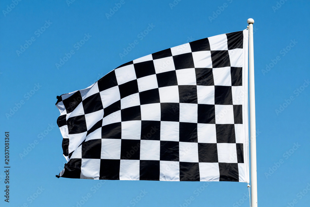 Obraz premium Black and white checkered flag flutters against a clear blue sky. Wind carries the flag with dynamic motion.